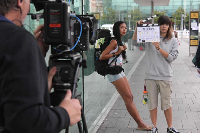 Un momento del rodaje de la película 'Blue Lips', con la actriz esperando con una mochila en la estación de autobuses mientras una integrante del equipo sujeta una claqueta. 